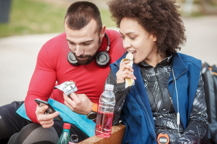 Young athletic man in sports clothing showing something on a smart phone to his partner while they are sitting next to each other in a public park Prinova